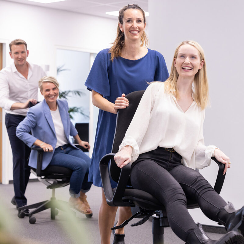 Schreibtischstuhl-Ralley im Büro, lebendiges und authentisches Imagefoto in Bewegung mit lachenden Menschen, Employer Branding Fotografie, echte Mitarbeitende, Laienmodelle