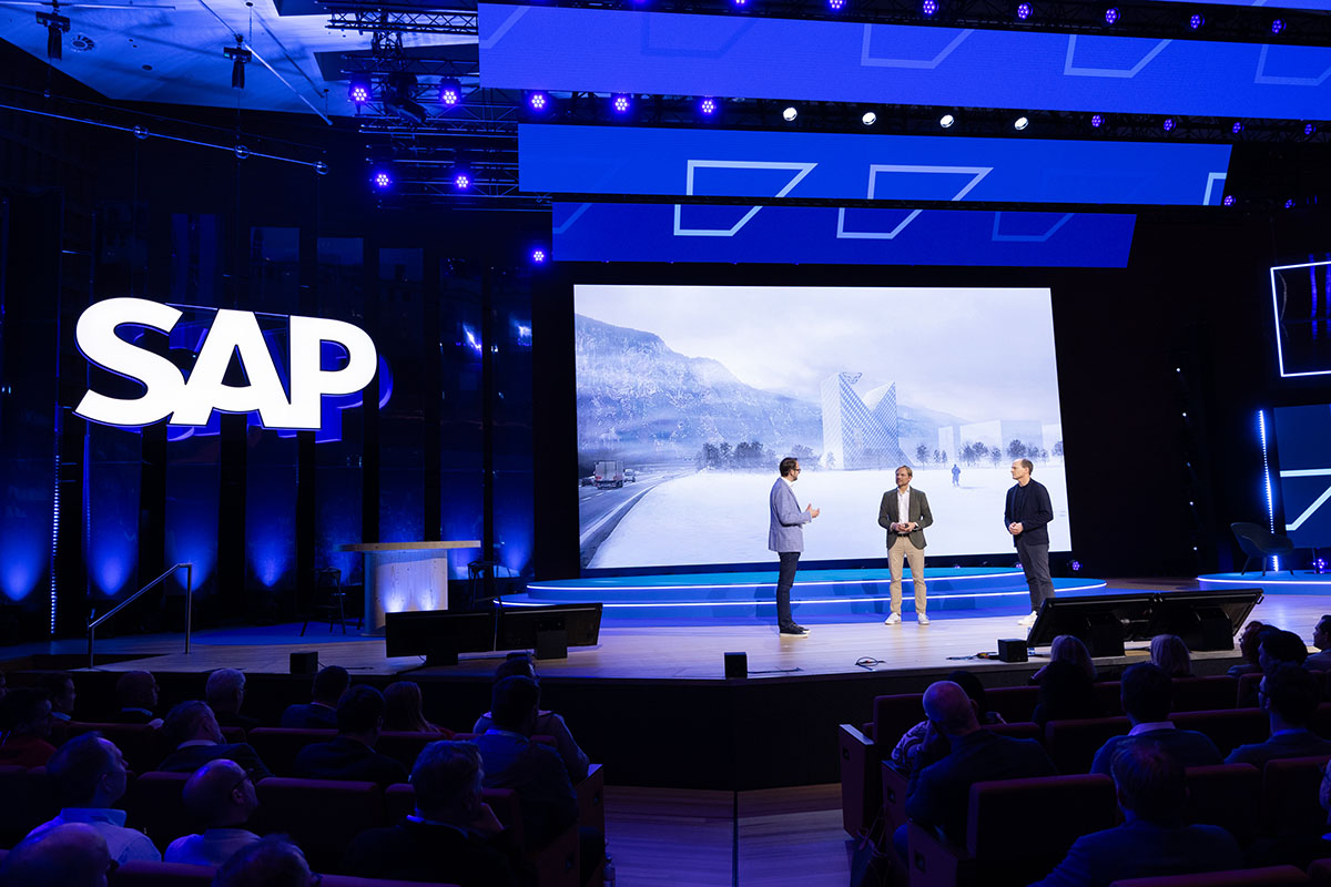 dunkelblaue große Bühne mit 3 Rednern vor einem großen monitor mit Schneelandschaft und SAP logo
